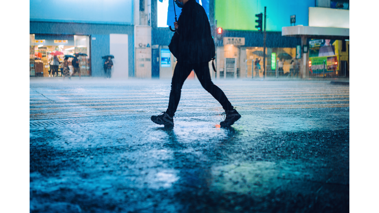 Silhouette of a man carrying umbrella walking in downtown city street against the reflection of glowing neon lights and city buildings in heavy rain at night