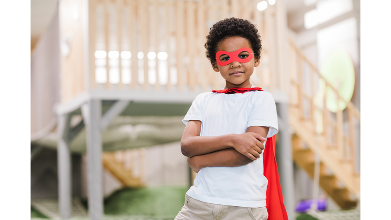 Cute African little boy in white casualwear and red mantle of superman