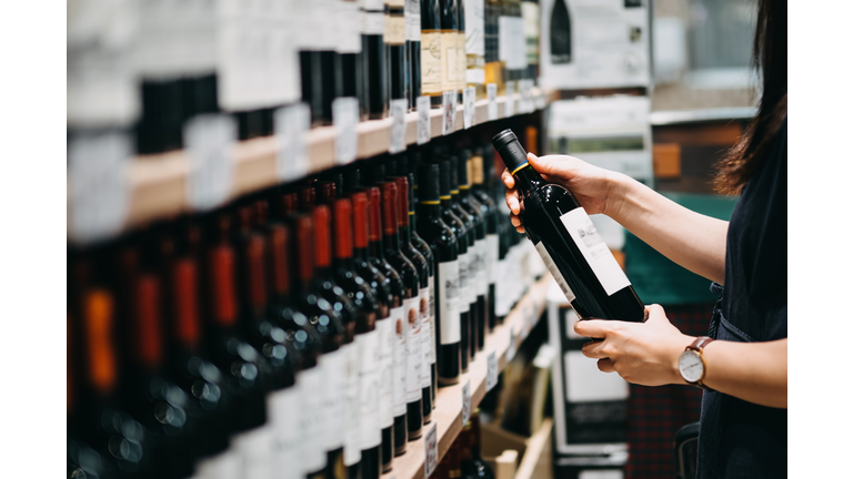 Close up of young Asian woman walking through supermarket aisle and choosing a bottle of red wine from the shelf in a supermarket