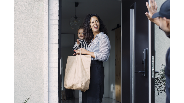Delivery man waving to smiling female customer carrying daughter while holding paper bag at doorway