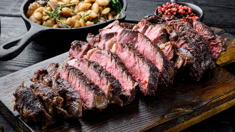 Sliced grilled meat steak Rib eye medium rare, on wooden serving board, with white beans and rosemary in cast iron pan, on black wooden table background
