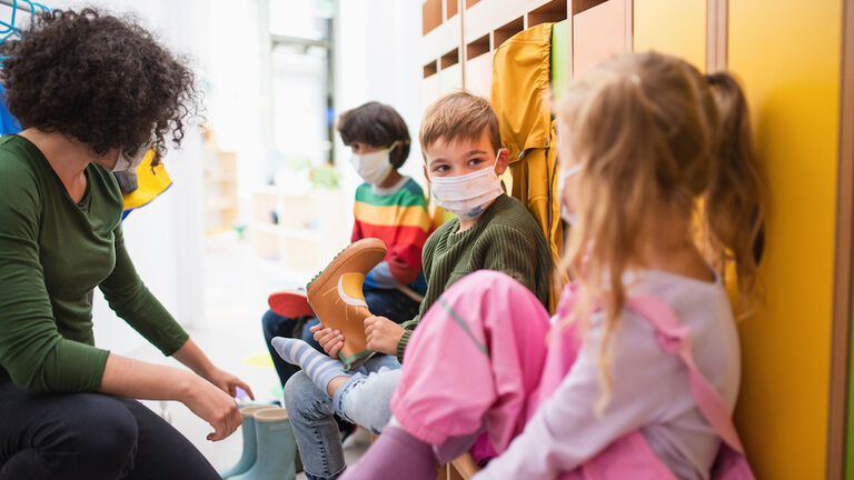 Pre school teacher helping children to put on shoes indoors in cloakroom at nursery, coronavirus concept.
