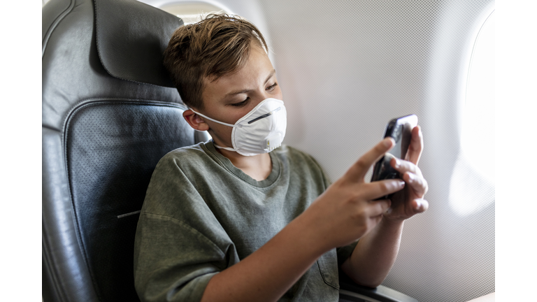 teenager boy is wearing his FFP3 protective mask while he is playing with his mobile phone in airplane