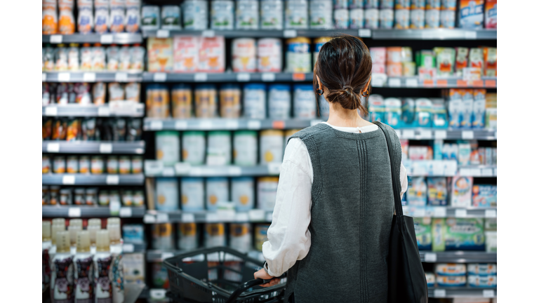 Rear view of young Asian mother with a shopping cart grocery shopping for baby products in a supermarket. She is standing in front of the baby product aisle and have no idea which product to choose from