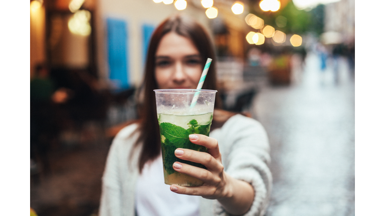 Street portrait of young woman holding a glass of mojito in hands. Blurred restaurant in background. Summer lifestyle, drinks concept.