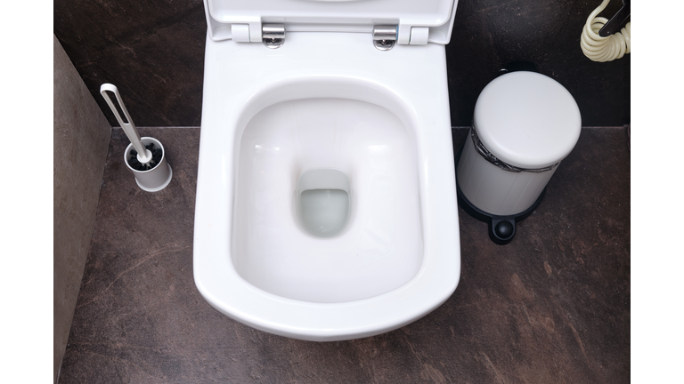 Clean toilet bowl with drain hole, top view. Toilet brush and trash can on black tiles