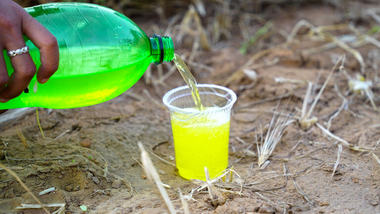 Human hand offering cold lemon juice in a plastic glass. Fruitee in plastic glass in a agriculture field.