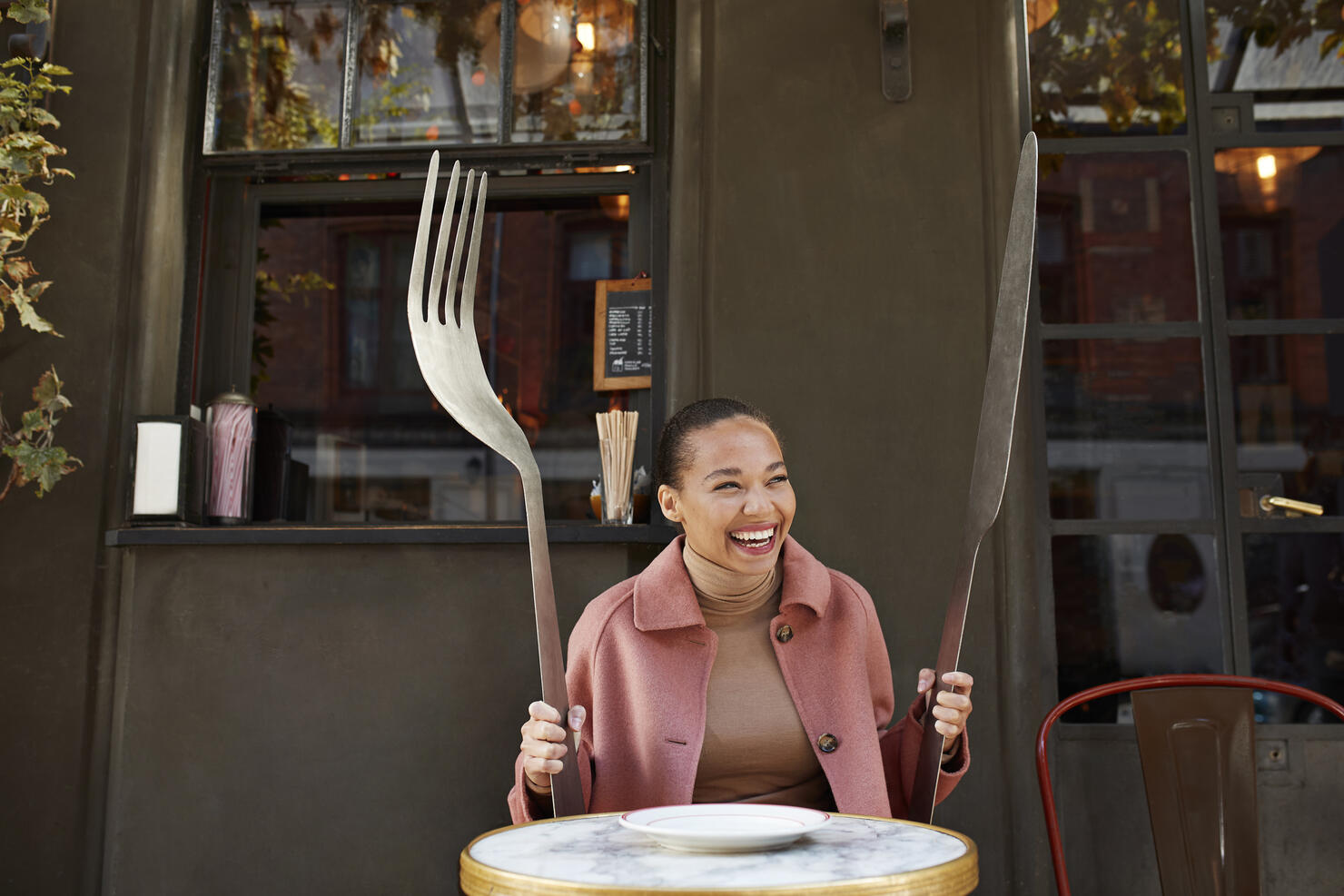 Happy woman holding large fork and table knife while sitting at sidewalk cafe