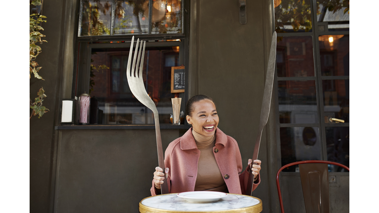 Happy woman holding large fork and table knife while sitting at sidewalk cafe