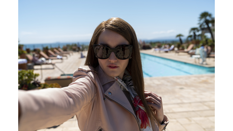 Brunette woman wearing large sunglasses and an overcoat standing next to a pool. 