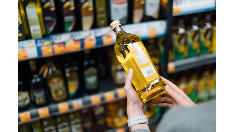 Close up of young woman grocery shopping in a supermarket. Standing by the aisle, holding a bottle of organic cooking oil, reading the nutritional label and checking ingredients at the back