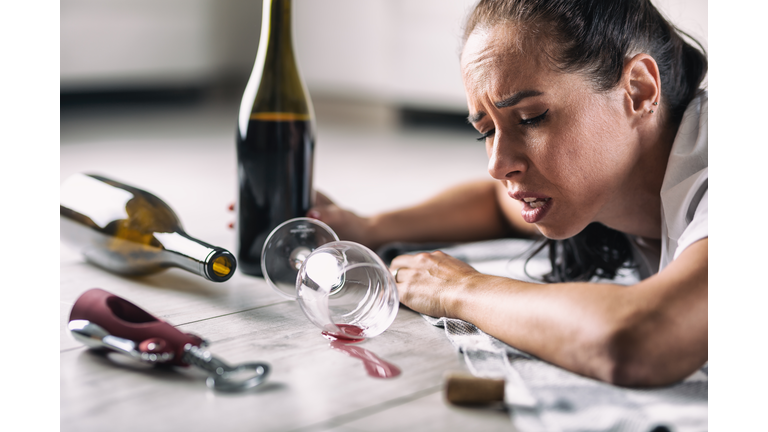 Hungover woman wakes up on the floor next to bottles of wine and spilled glass.