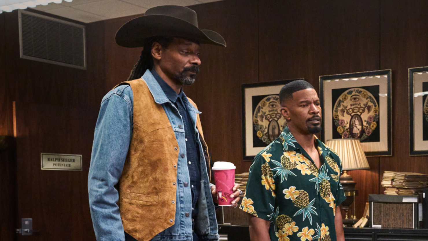 Big John Elliot (Snoop Dogg) wears a tan cowboy vest and ten-gallon hat and sips coffee while standing next to Bud (Jamie Fox), who is wearing a pineapple button up shirt, in Day Shift.
