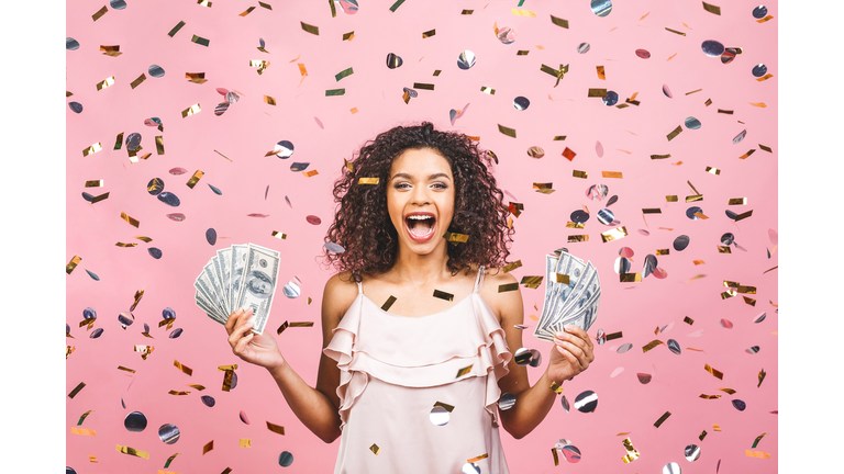 Black afro american girl won money. Happy young woman holding dollar currency satisfied isolated over pink background with confetti.