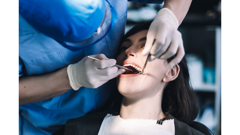 Un dentista con bata azul, guantes de látex, gafas transparentes y mascarilla limpia los dientes de un paciente con una herramienta metálica.