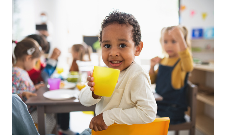 Pre school multiracial boy having meal with children indoors at nursery school, looking at camera.