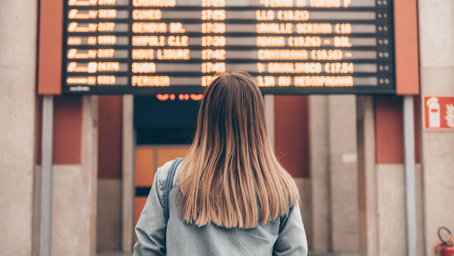 A young woman at a railway station or at the airport looks at the smartphone screen against the background of the arrival and departure board