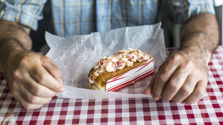 A man about to eat a lobster roll on a table with a red cheked tableclothes