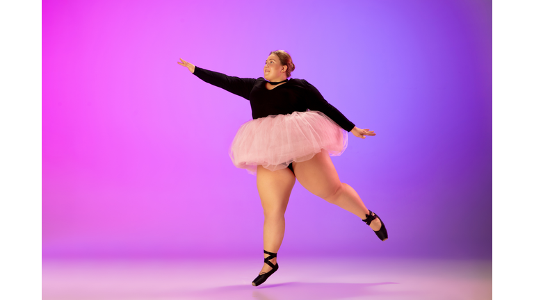 Beautiful caucasian plus size model practicing ballet dance on gradient purple-pink studio background in neon light