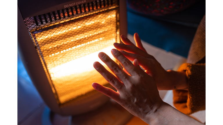 A woman warms her hands on an electric heater at home in the