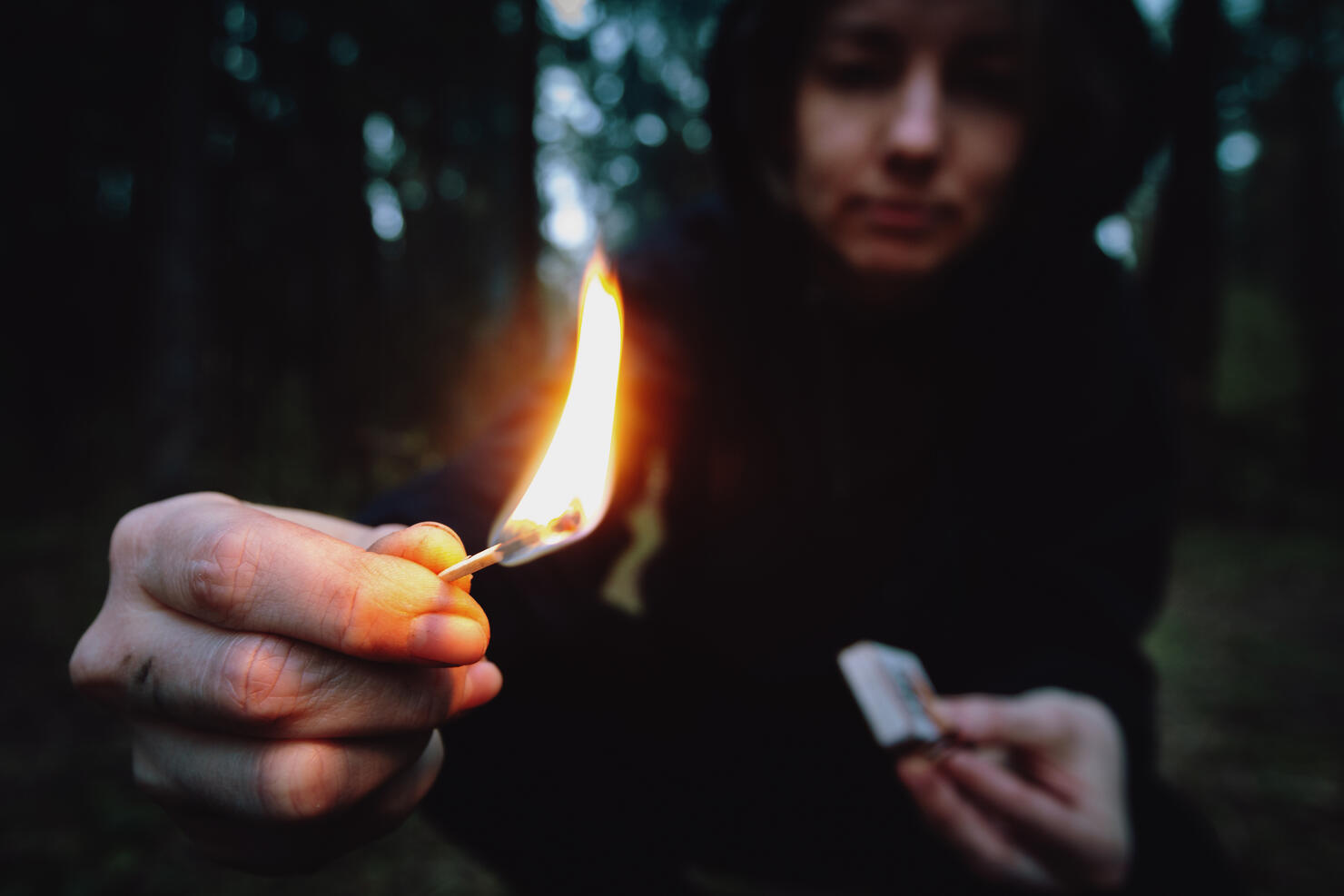 A teenager in a black hood holding a burning match in a dark forest at night. Arsonist damages the environment. Forest fire danger, flame in the hands of a hooligan
