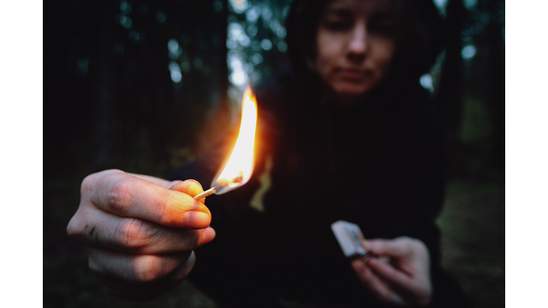 A teenager in a black hood holding a burning match in a dark forest at night. Arsonist damages the environment. Forest fire danger, flame in the hands of a hooligan