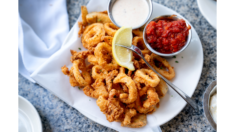 A Delicious Plate Of Deep Fried Calamari, Squid Fried To Perfection Cajun Style, Served With A Side Of Freshly Made Garlic Aioli Sauce And Cocktail Sauce, Fresh Lemon Wedges On The Side
