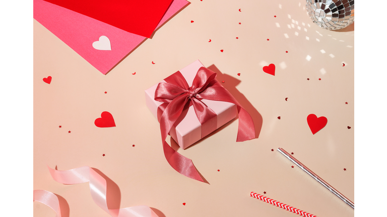 Valentine day composition concept: white gift box with magenta colored bow, red hearts confetti and pink paper on pastel background. High angle view, flat lay