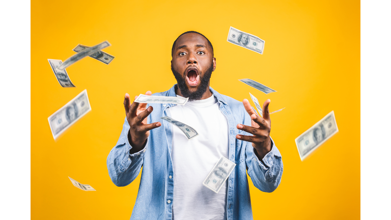 Portrait of a happy young afro american man throwing out money banknotes isolated over yellow background.