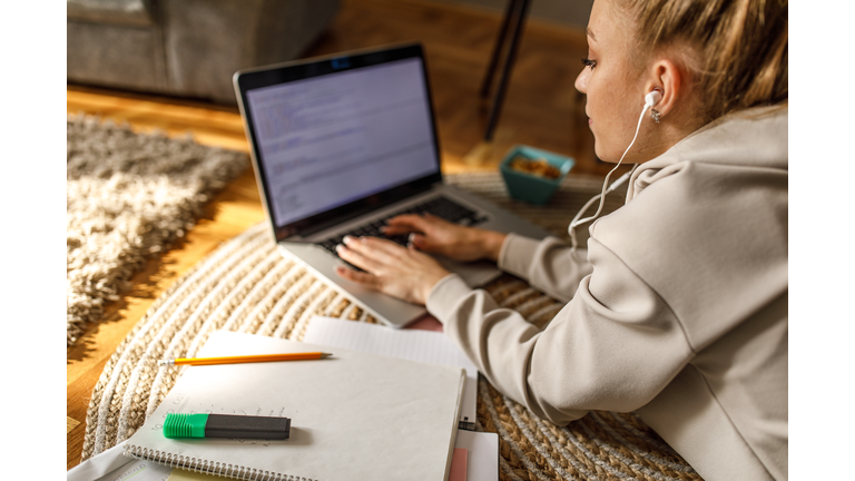 Teenage girl lying on the floor, tying an essay on laptop for homework