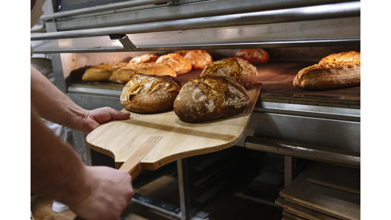 Male chef with pizza peel removing baked bread from oven at bakery