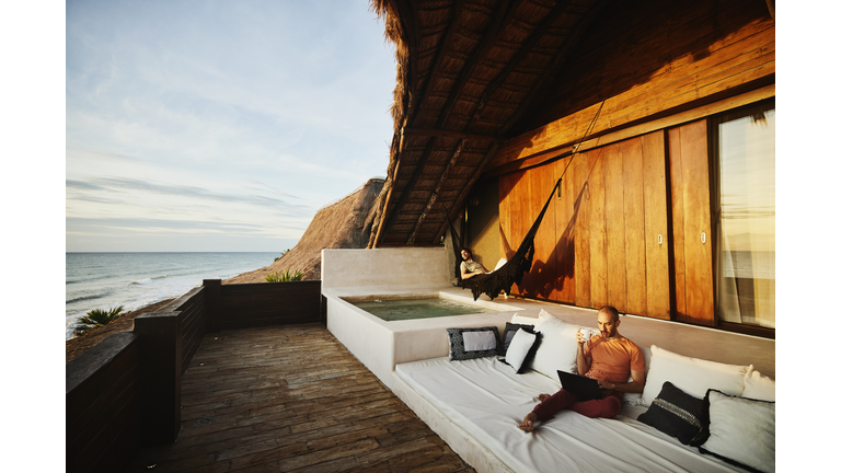 Wide shot of man drinking coffee and working on laptop while partner relaxes in hammock on deck of luxury tropical beach villa at sunrise