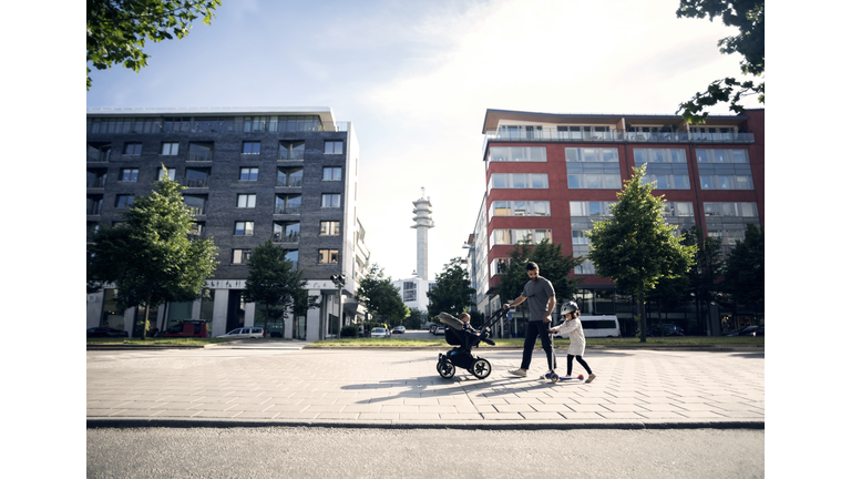Father walking with baby stroller looking at daughter riding push scooter in city