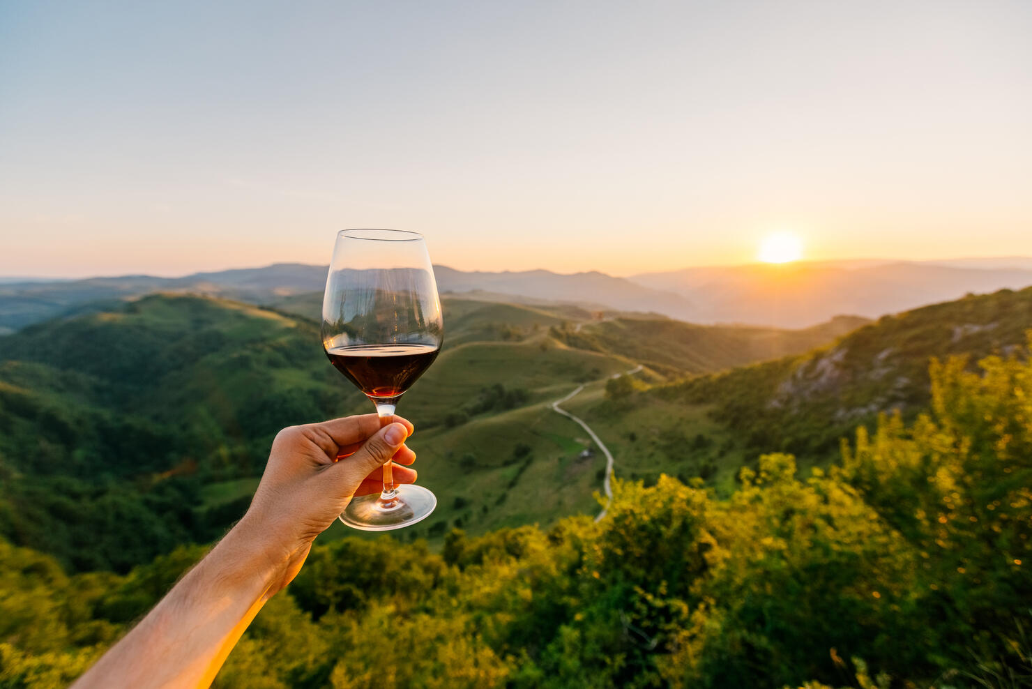 Man holding a glass of red wine surrounded by hills and mountains at sunset, personal perspective POV