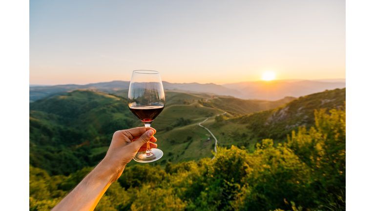 Man holding a glass of red wine surrounded by hills and mountains at sunset, personal perspective POV