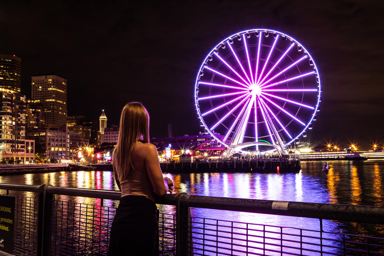 Woman Standing Alone Looking at Seattle Washington at Night The Great Seattle Wheel Lit Up Reflecting on Pier 69 Harbor in the Pacific Ocean Long Exposure Photo with City Lights in the Background