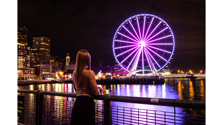 Woman Standing Alone Looking at Seattle Washington at Night The Great Seattle Wheel Lit Up Reflecting on Pier 69 Harbor in the Pacific Ocean Long Exposure Photo with City Lights in the Background