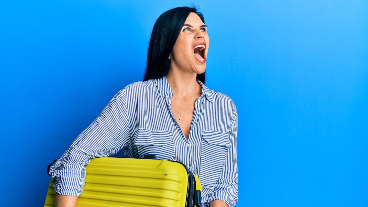 Young caucasian woman holding cabin bag angry and mad screaming frustrated and furious, shouting with anger. rage and aggressive concept.