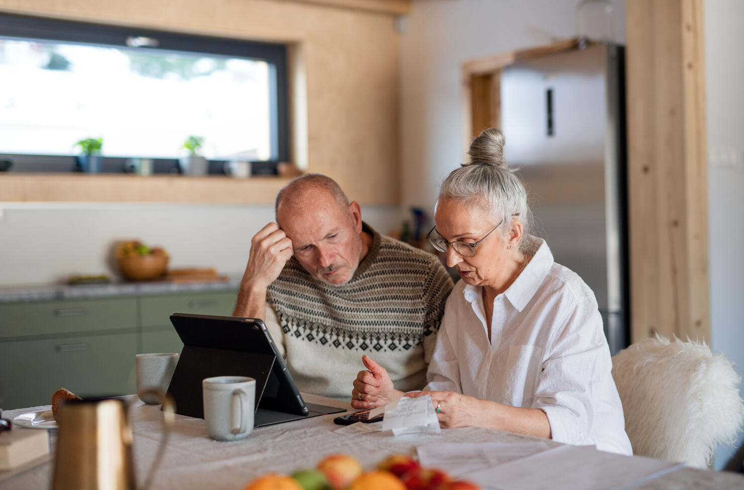 Senior couple sitting at the kitchen table looking at digital tablet and recalculating their expenses.