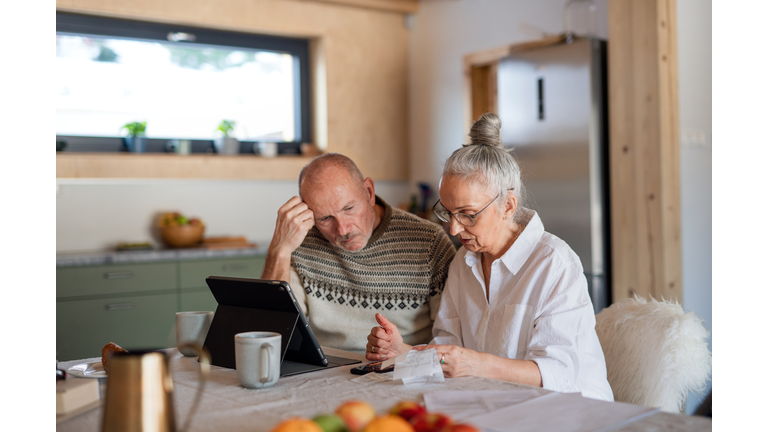 Senior couple sitting at the kitchen table looking at digital tablet and recalculating their expenses.