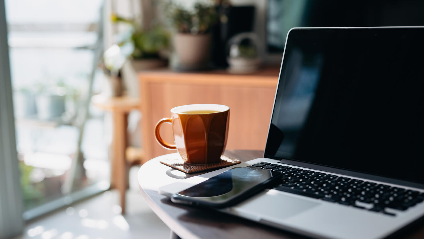 Cropped shot of wooden coffee table with laptop, smartphone and a cup of tea in the living room at home by the window against beautiful sunlight