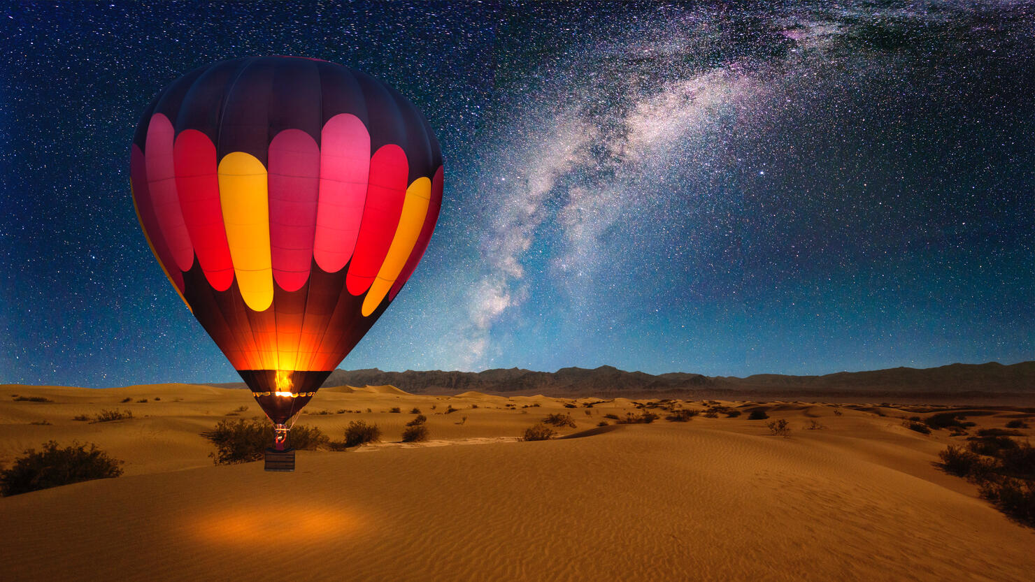 A majestic hot air balloon soars under the stars of the Milky Way, over the desert - Mesquite Dunes of Death Valley National Park. Moonlight provides luminosity showing the patterns and shapes of the desert landscape.