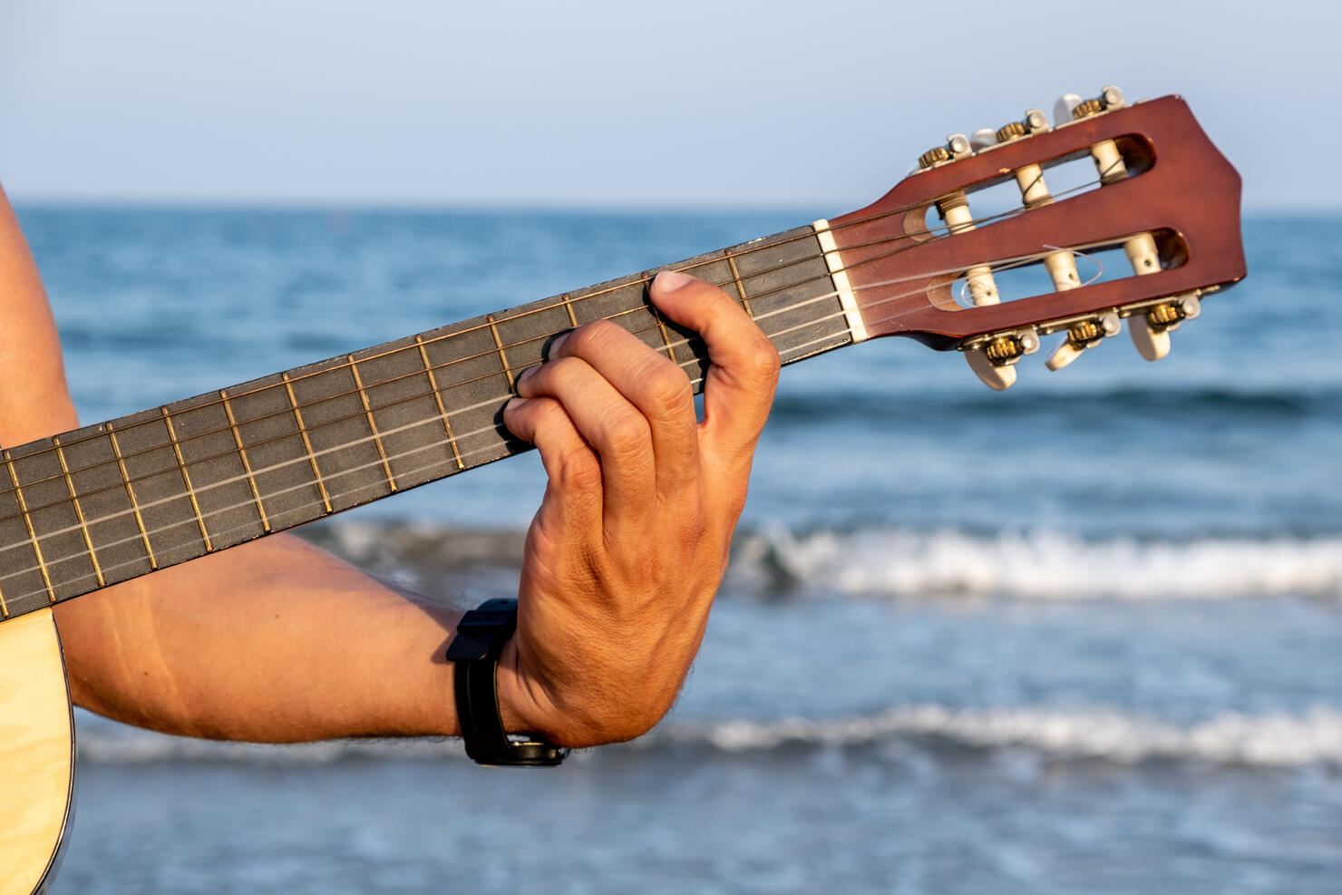 A man's hand holds a Spanish guitar neck with the sea in the background.
