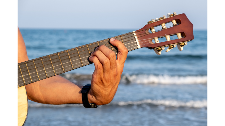 A man's hand holds a Spanish guitar neck with the sea in the background.