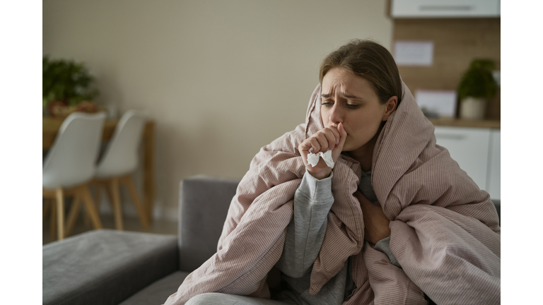 Caucasian young woman coughing and sitting under the duvet at home