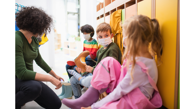 Pre school teacher helping children to put on shoes indoors in cloakroom at nursery, coronavirus concept.