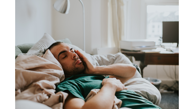 A man wakes up on a sofa, surrounded by sheets and duvets and pillows. He rubs his eyes and face as he tries to rouse himself.