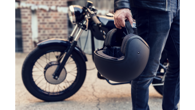 Unrecognizable motorcyclist carries his motorcycle helmet in one hand. In the background is his parked motorcycle