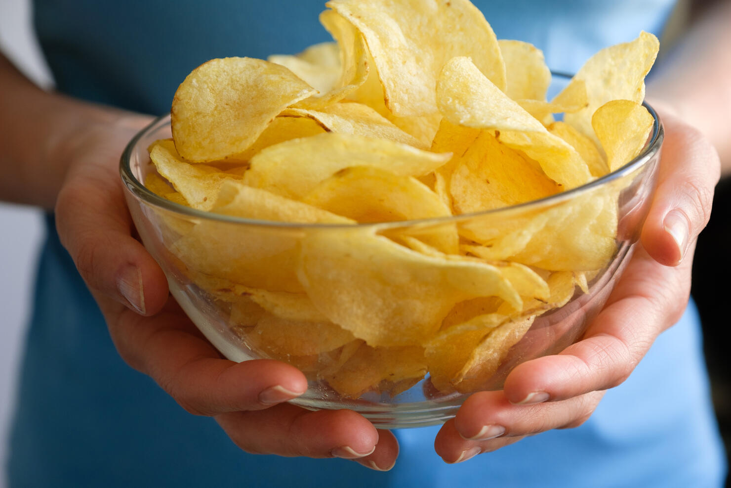 Crispy fried fatty potato chips in a glass bowl or plate, on a white background or table. Chips in the hands of a woman or a teenage girl, she eats them. The concept of an unhealthy diet and lifestyle, the accumulation of excess weight.