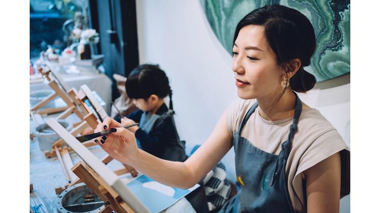 Young Asian mother and little daughter having a painting class together. Painting on a canvas with paintbrush and colourful acrylic paints. Having a relaxing afternoon